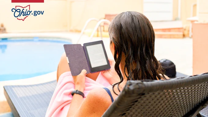 A woman sitting beside a swimming pool and reading a book on an e-reader.