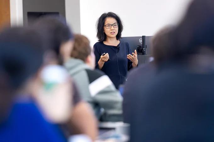 A female scientist, wearing a microphone and holding a remote control for a slide show, presents her scientific findings to a large group of fellow researchers inside a modern lecture hall on the campus of a university.