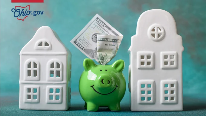 A bright green ceramic piggy bank with a one-hundred dollar bill sticking out sits next to white ceramic houses.