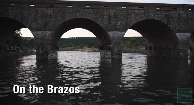 Video still of stone bridge over river. Title on screen says On the Brazos