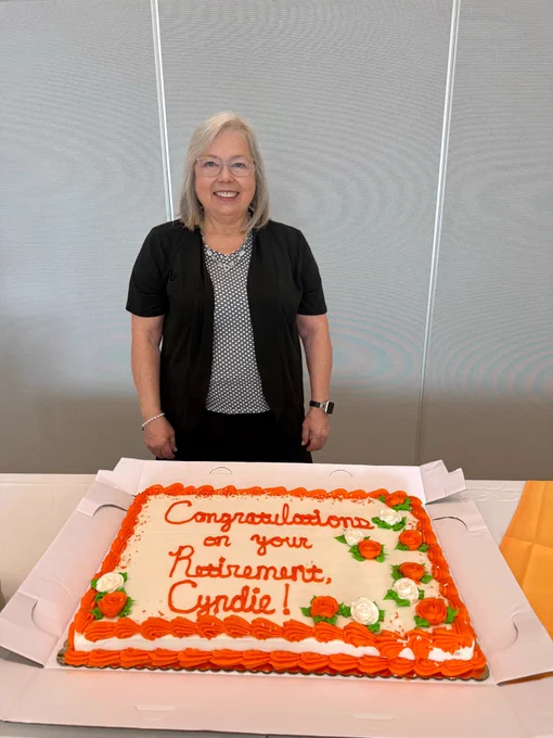 A woman with shoulder-length gray hair wearing a black jacket, black skirt, & black & white designed shirt standing in front of a large sheet cake. Cake is white frosting with orange piping and white and orange frosting. The words on the cake are "Congratulations on your retirement, Cyndie!"