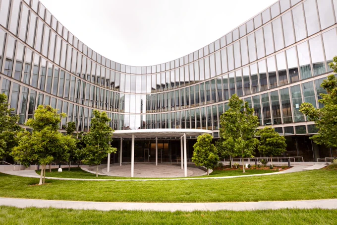 Curved glass exterior and south courtyard of the Barbara and Gerson Bakar Gateway, home of the UC Berkeley College of Computing, Data Science, and Society, featuring a shade canopy, landscaped trees and tiered lawn area.