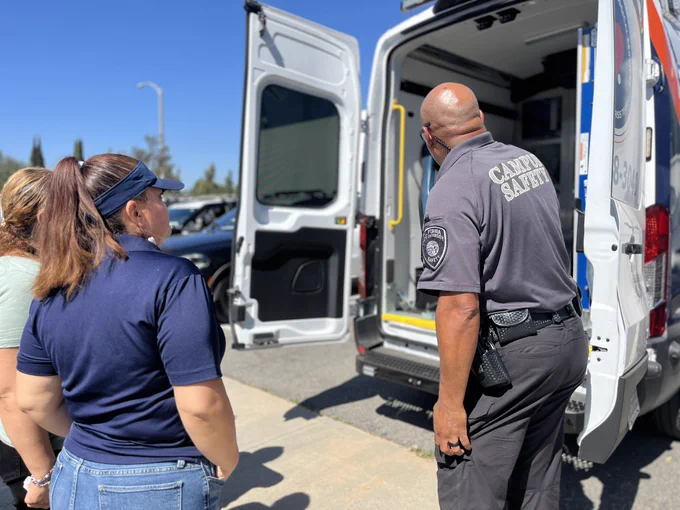 Three people stand behind an open campus MCT van on a sunny day. A campus safety officer in uniform faces the van’s interior while two casually dressed individuals stand nearby, looking inside. The van’s rear doors are open, revealing steps and equipment, with parked cars and a clear blue sky in the background.