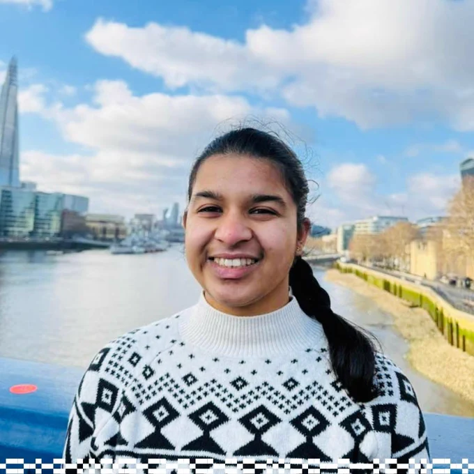A person is standing on a bridge with a cityscape in the background. The Shard is visible among the skyline. The river flows alongside modern buildings under a partly cloudy sky. The person is wearing a patterned sweater and smiling.