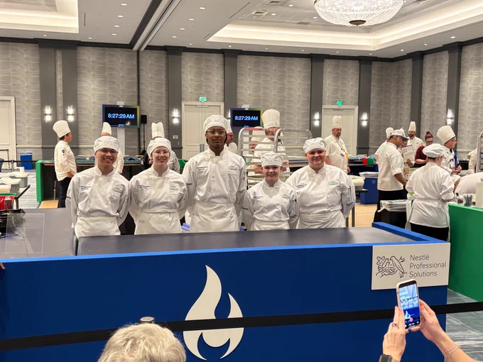 The Elk River High School team stands together wearing white competition chef hats, jackets and aprons in their competition booth. 