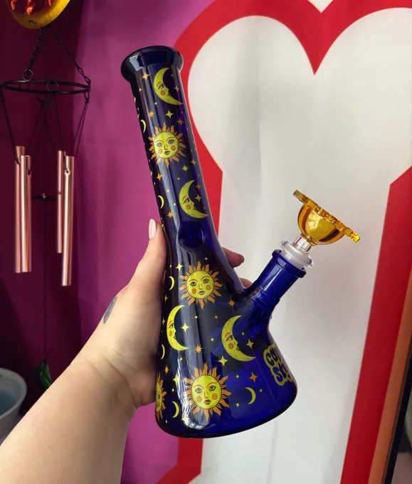 a picture of a dark blue bong/ water pipe that’s covered in 90’s themed sun and moon decals 

there is also a sun shaped bowl piece that sits in the downstem 

in the background a lesbian flag can be seen, as well as a small bit of Bug’s sun and moon windchime