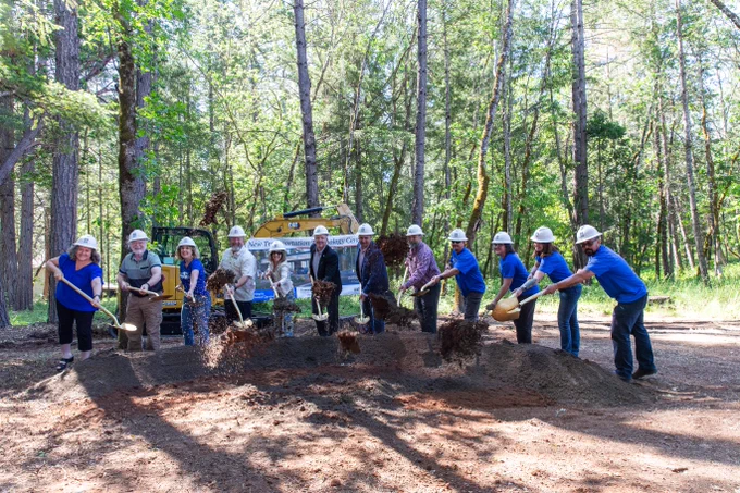 A group of people from RCC and the local community wearing hard hats and holding shovels participate in a groundbreaking ceremony in a forested area at RCC's Redwood Campus. A Caterpillar construction vehicle is visible in the background.