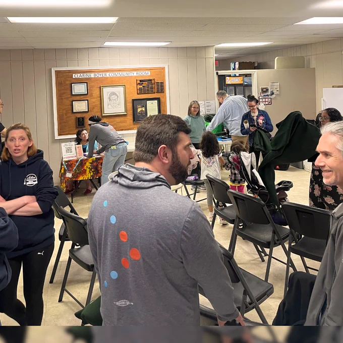 Adults and kids are gathered in a room talking to eachother and socializing. You can see the room is labeled "Clarine Boyer Community Room." 
