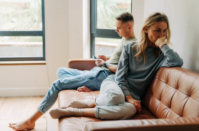 Two people sit on opposite ends of a brown sofa in a room with large windows. One person looks at a phone, while the other gazes thoughtfully into the distance. Both appear relaxed, wearing casual clothes.