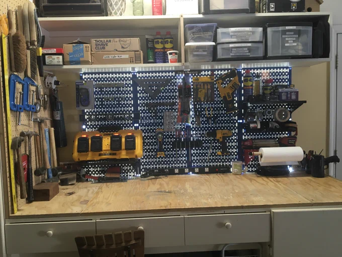 Organized garage workshop featuring a Wall Control metal pegboard system mounted above a wooden workbench, holding power tools, hand tools, chargers, measuring tools, and accessories. Storage bins and shelves are mounted above the pegboard, with additional hand tools hanging on a side pegboard wall. Photo courtesy of customer Paul.