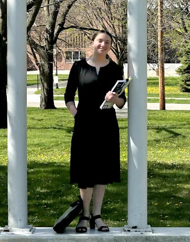 UW-Madison Badger Precollege student standing in front of two white columns wearing a black dress and holding a book in her arm.