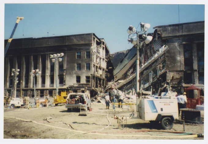A color photograph of the Pentagon building after the 9/11 terrorist attacks. The center part of the building in the photo is collapsed, the interior is visible at an angle, and the outside walls of each side are blackened from smoke and fire. There is the arm of a crane in the left side of the photo. Pop-up flood lights are positioned in different spots in front of the building and there is a truck with the trunk open in the center of the photo. People with hard hats are scattered throughout .