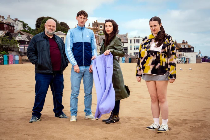 Group image. Four people in differing attire stand on a sandy beach. A man on the left wears a leather jacket, jeans and trainers, while a younger man beside him wears a tracksuit jacket and jeans. The woman next to him wears a long coat and holds a purple pair of jogging bottoms. Stood slightly apart from the group is a young woman wearing a fleece and a long t-shirt. 