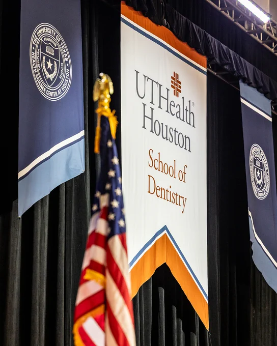 A stage is set for a formal event with an American flag in the foreground. A large central banner reads “UTHealth Houston School of Dentistry” with the UTHealth logo above it, flanked by two blue banners displaying the seal of The University of Texas Health Science Center at Houston.