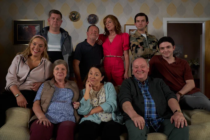 The cast of Two Doors Down series 7 sit on and stand around the sofa in Beth and Eric's living room