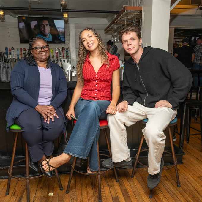 Three attendees sit on barstools smiling at the camera during a Stony Brook GOLD alumni happy hour at a restaurant bar area.