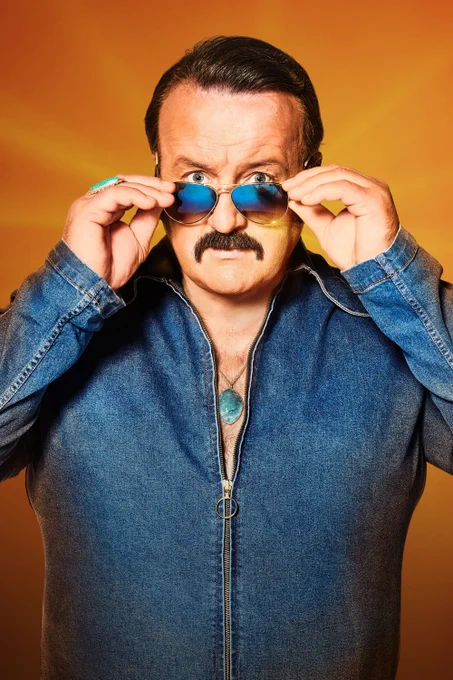 A man wearing a denim zipped jacket, aviator sunglasses and a blue pendant necklace. He lowers his sunglasses with both hands to look directly to the camera. 
