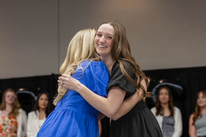 Two people are embracing at an event. One is wearing a blue outfit, and the other is in black. Several others are sitting and observing in the background.