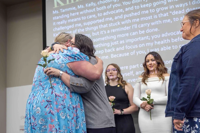 Two women embrace on a stage. One holds a white rose. Several women surround them, looking on. A screen with text appears in the background.