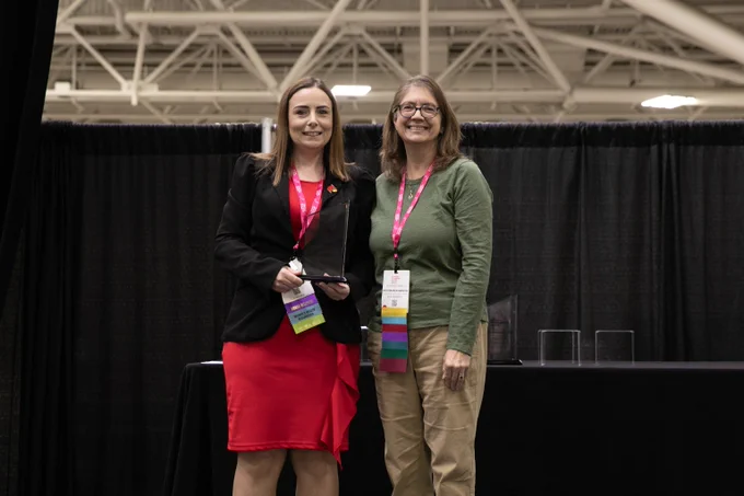 Two women standing side by side at an indoor event, one holding a clear award plaque, both wearing event badges and lanyards.