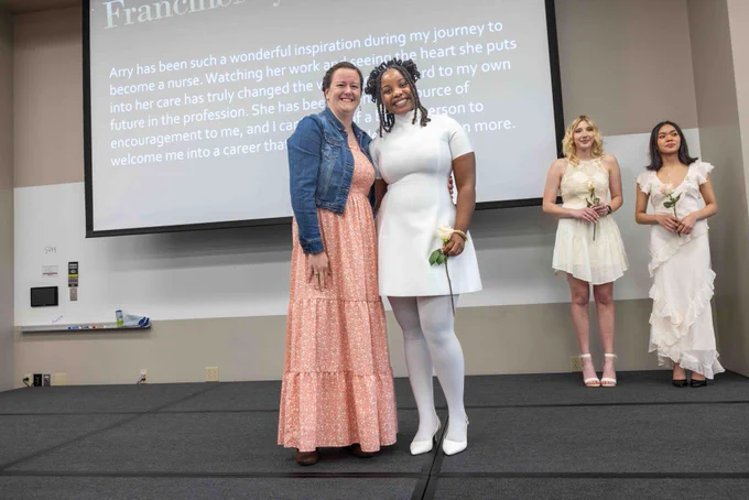 Two women smile at the camera, standing on a stage. One wears a long pink dress, and the other wears a short white dress and holds a white rose. A screen with text appears behind them. 