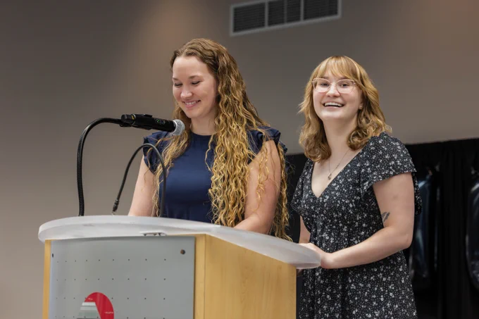 Two people smiling and standing at a podium. They appear to be speaking into a microphone. The podium has a logo featuring a red circle.
