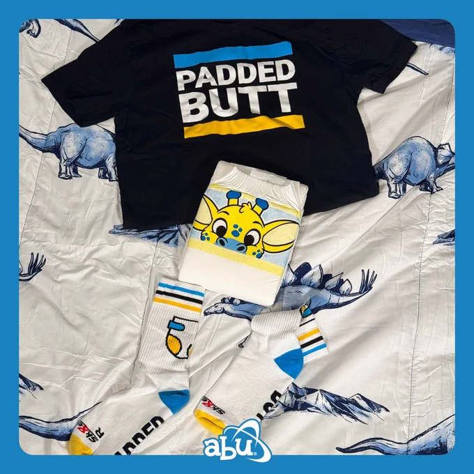 Photo of a set of socks, an adult diaper, and a tee shirt laid out on a bed. The Shirt and socks are Sk8erBoy's "Padded Butt" line. Featuring bold blue and yellow coloring with a diaper graphic on the socks and tee with the words "Padded Butt" on both tee and socks. The adult diaper is an ABU PeekABU. A white diaper with cartoon character animals on the landing zones.