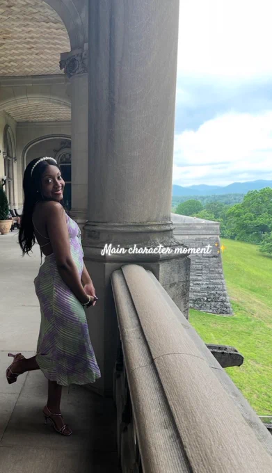 Woman in a pastel purple and green dress posing on a stone balcony at the Biltmore Estate overlooking rolling green hills and mountains.