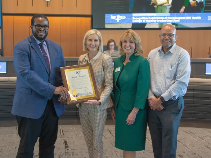 Four individuals are standing together indoors, dressed in business attire. One person is holding a framed certificate. Behind them is a sign that reads "Fort Worth" and mentions a recognition event for the UNTH Health College of Nursing graduating class.