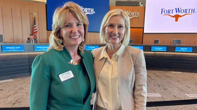 Two individuals are smiling at a Fort Worth City Hall Council Chambers. They stand in front of a long table with nameplates and multiple Fort Worth logos are visible on screens and a wall. An American flag is displayed in the background.
