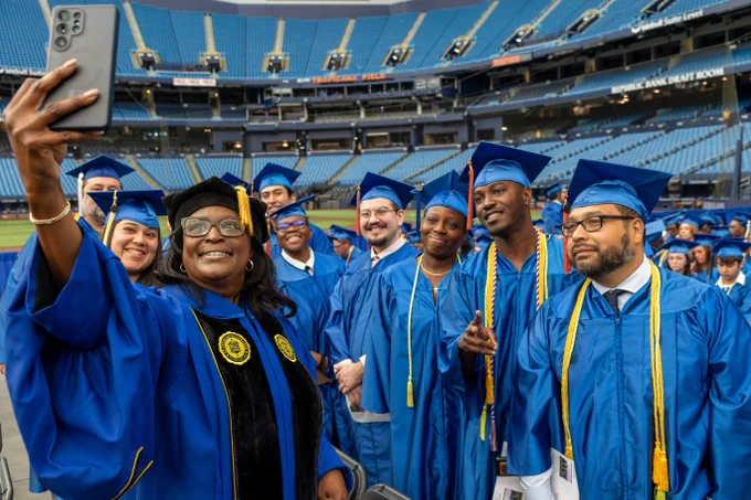 A group of graduates wearing blue gowns and caps gather in a stadium to take a selfie. SPC President Dr. Tonjua Williams wears academic attire and holds the phone for the photo.