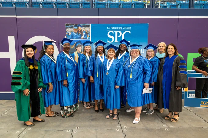 A group of graduates and faculty in gowns and caps stand together, smiling, in front of a sign for St. Petersburg College.