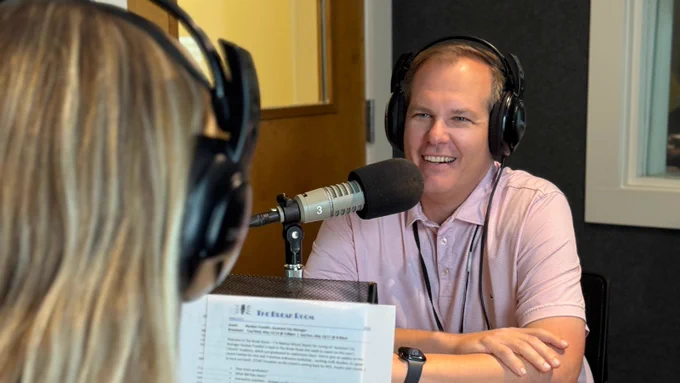A person is speaking into a microphone in a recording studio, wearing headphones and a light pink shirt. Another person, also in headphones, faces away from the camera, holding a document. The atmosphere suggests a radio or podcast recording session.