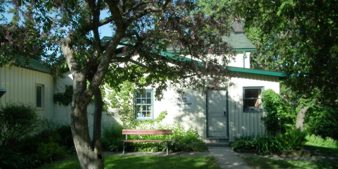 A white building surrounded by greenery and trees with a wooden bench in front.