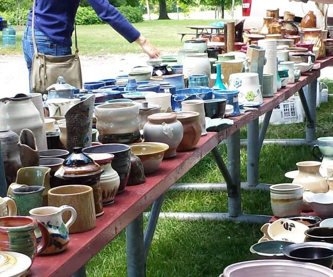 A person browses a display of handmade ceramic pottery on outdoor tables.