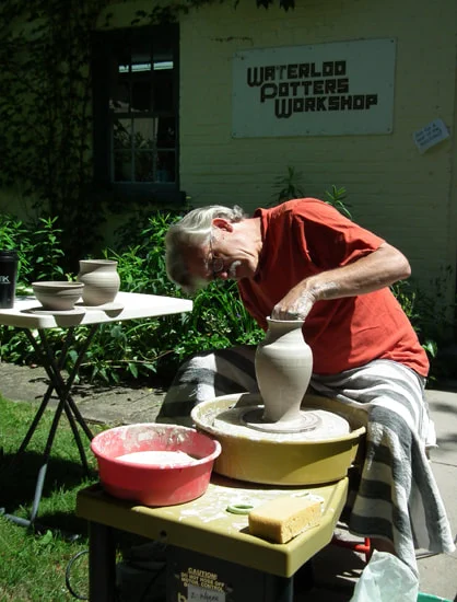A person seated outdoors is focused on shaping a clay pot on a pottery wheel with a sign on the wall behind that says "Waterloo Potters Workshop".