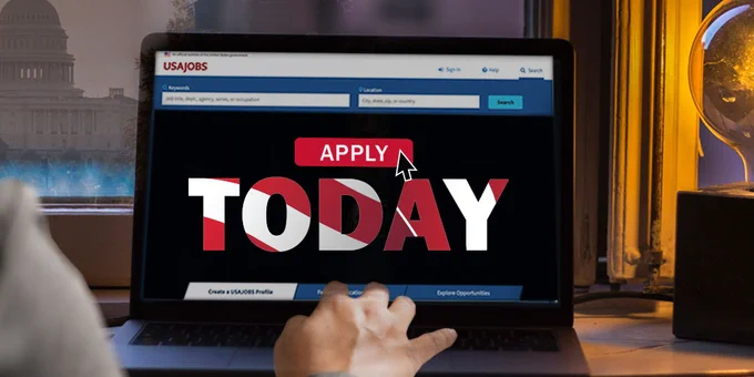 A person sitting at a desk with a laptop displaying the USAJOBS website homepage. The screen shows a large "Apply Today" button.