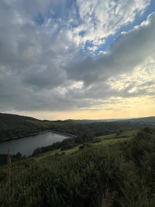 A scenic landscape of a rolling green hill overlooking a large reservoir, under a sky filled with clouds and soft sunlight breaking through.