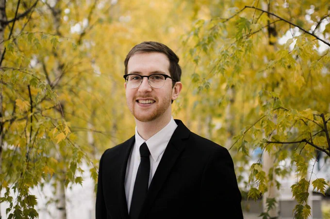 Headshot of Thomas Russel, standing in front of green foliage.