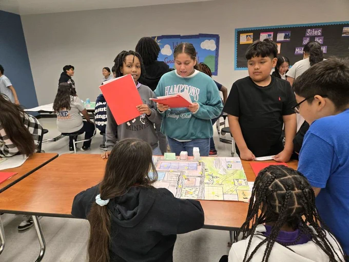 A group of students gather around a table with a map, discussing and looking at books.