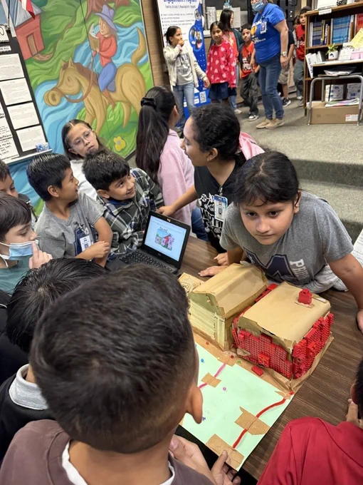 Children examine a cardboard house on a table in a classroom, some looking at a laptop.