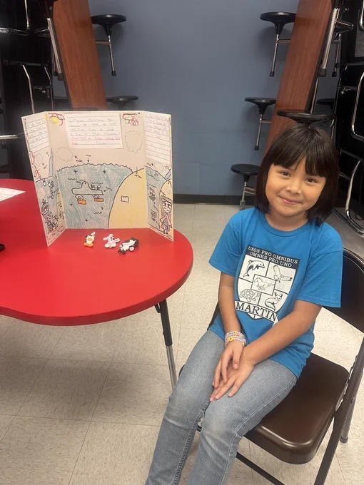 Child seated at a round table, smiling, with a display board behind them on the table.