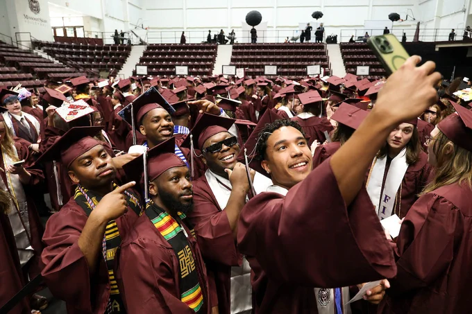 A group of graduates in maroon caps and gowns take a selfie together at a commencement ceremony. The background features bleachers filled with more seated graduates.