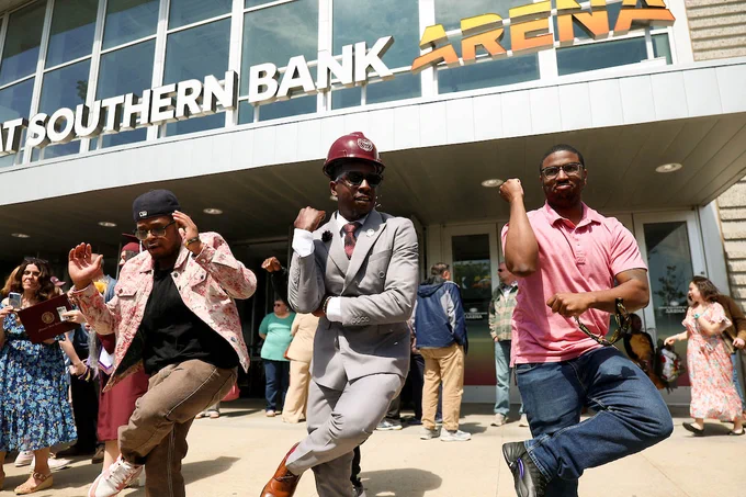 Three people are dancing energetically in front of Great Southern Bank Arena. The person in the center is wearing a suit and maroon hat, while the others wear casual clothing. A crowd watches in the background.
