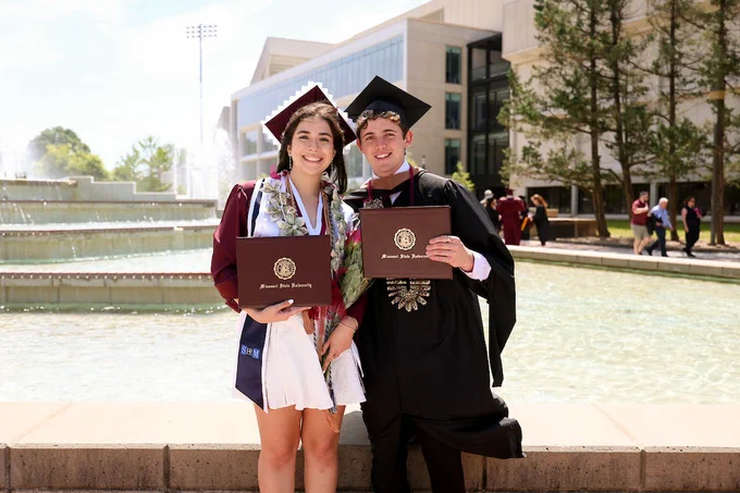 Two people in graduation attire stand smiling, each holding a diploma from Missouri State University. They are positioned in front of a fountain with a large building in the background.