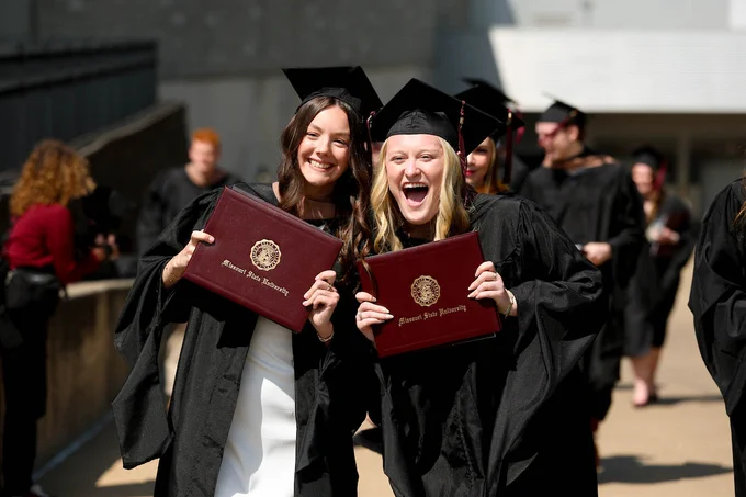 Two graduates in caps and gowns hold diplomas from Missouri State University, smiling and celebrating. Others in similar attire are in the background.