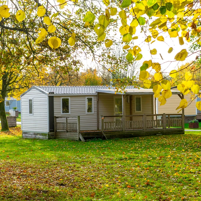 A mobile home is situated in a grassy area surrounded by autumn trees with yellow leaves. The foliage partially frames the image, creating a serene, natural setting.