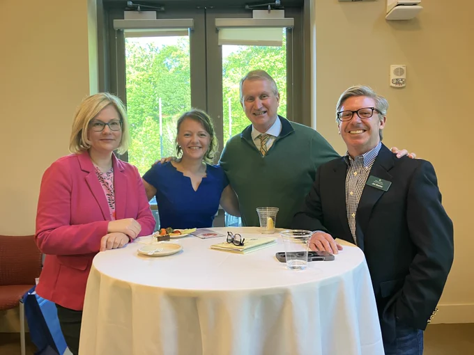 Four people stand together around a tall, round table at an indoor university reception. They face the camera in business or business-casual attire, with light refreshments on the table. Large windows behind them let in natural light, creating a warm, social atmosphere.