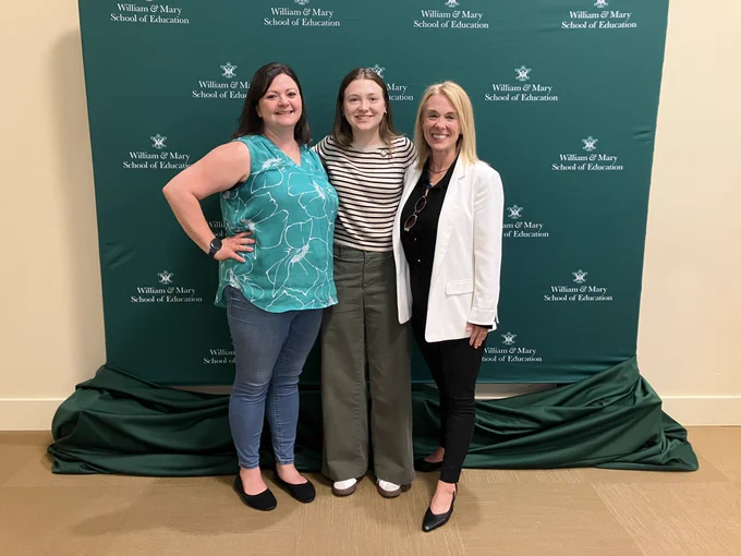 Three people stand side by side and pose for a photo in front of a green backdrop labeled ‘William & Mary School of Education.’ They wear business-casual attire and smile toward the camera, creating a professional and celebratory tone.