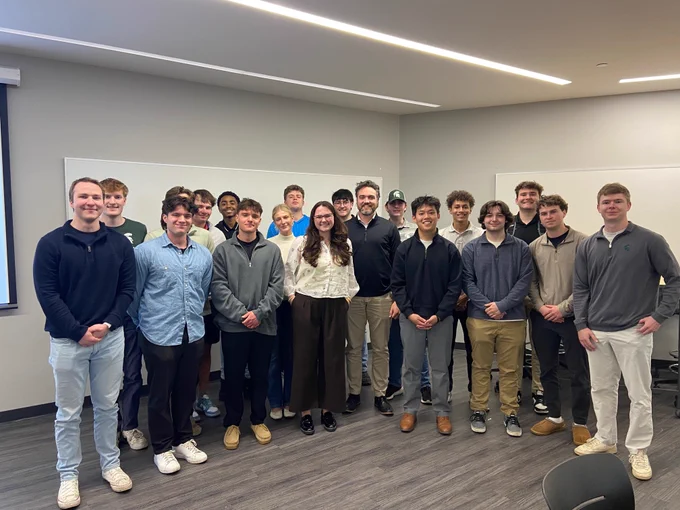 A group of 20 young adults and one older adult pose in a classroom. Most of the group are white males, and one is a white female. The older adult is a white male with a beard.